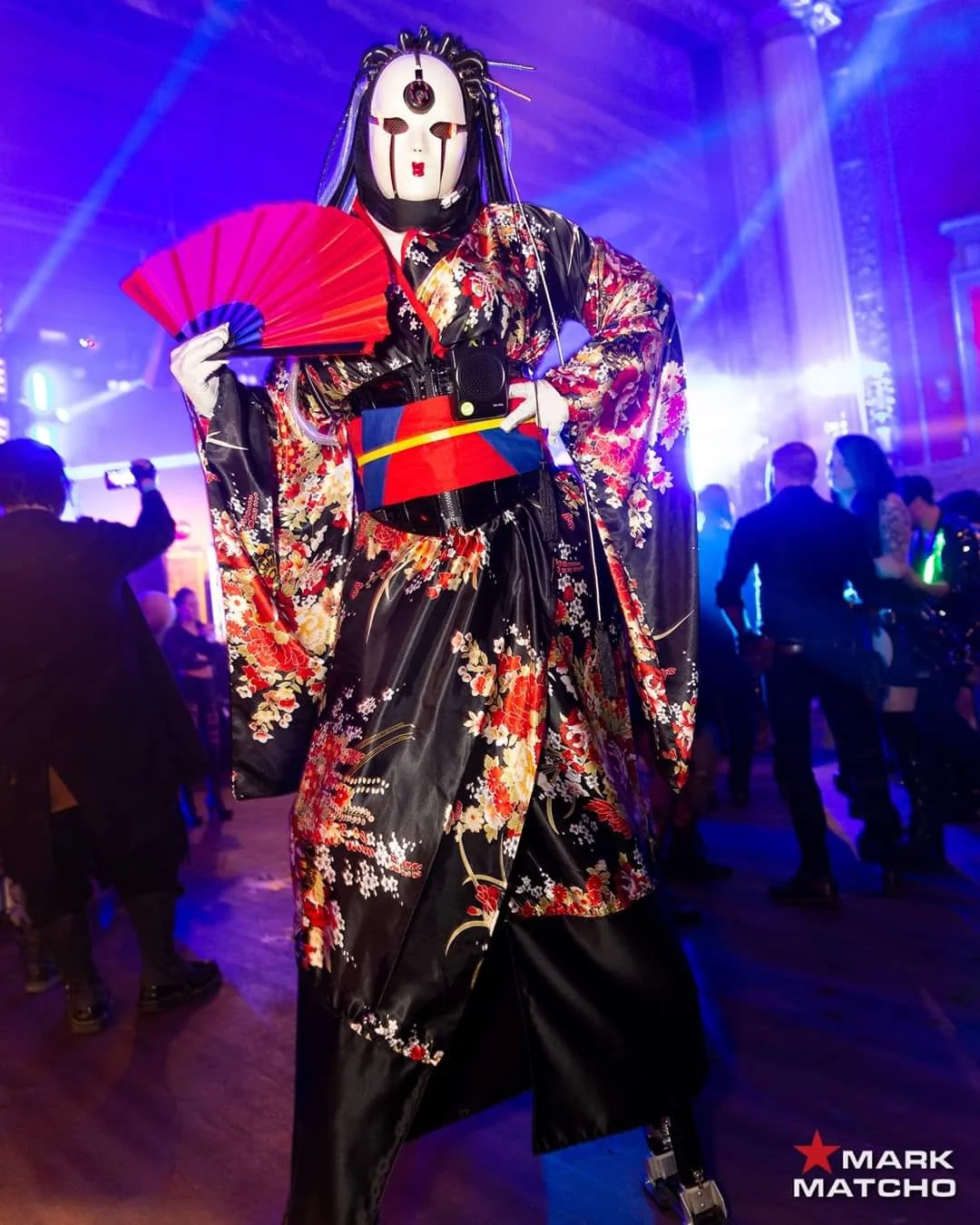 ROSHIE performing in a floral robe with a red fan under purple light